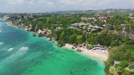drone view of padang padang’s turquoise cove below uluwatu cliffs: reef lines, white sand, sunbathers and clifftop resorts under a bright tropical sky, left-orbit aerial shot
