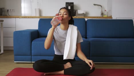 Mujer-Afroamericana-Bebiendo-Agua-Después-De-Hacer-Ejercicio-En-Casa-En-Una-Estera-De-Yoga