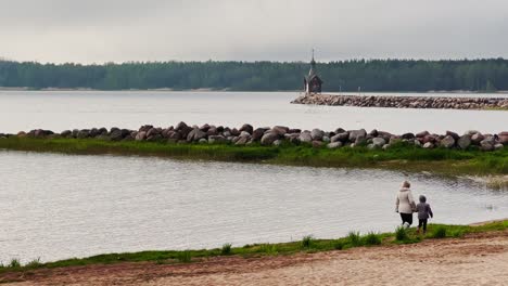 mother with child walks on the beach against the background of old chapel stands on a stone promontory on the water in leningrad region russia at cloudy weather, the forest in the background