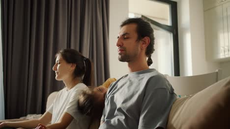 A-brunette-man-in-a-gray-T-shirt-and-his-wife,-a-brunette-girl-in-a-white-T-shirt,-meditate-while-sitting-on-the-sofa-and-their-little-brunette-daughter-in-a-yellow-dress-distracts-them-and-draws-attention-to-herself-in-a-modern-apartment