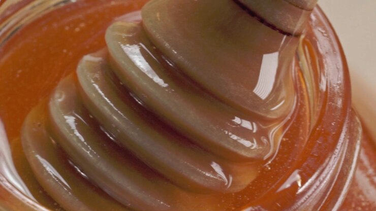Macro close up of honey dipper coated with fresh golden honey inside glass jar. Thick natural honey flows around wooden dipper with glossy texture