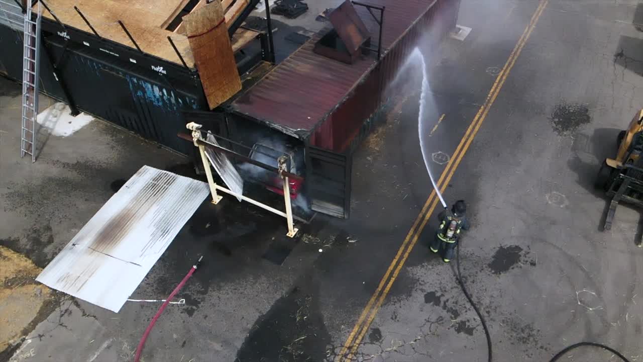 rallentatore drone riprese aeree del vigile del fuoco in giacca casco completo equipaggiamento estinguere il fuoco nel camion in contenitore di metallo panning su acqua zampillante e servizi di emergenza tubo flessibile