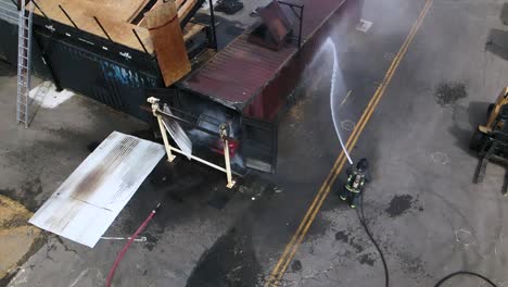 imágenes aéreas de drones en cámara lenta de un bombero con una chaqueta de casco de equipo completo extinguiendo el fuego en un camión en un contenedor de metal que se desplaza sobre los servicios de emergencia de agua y mangueras