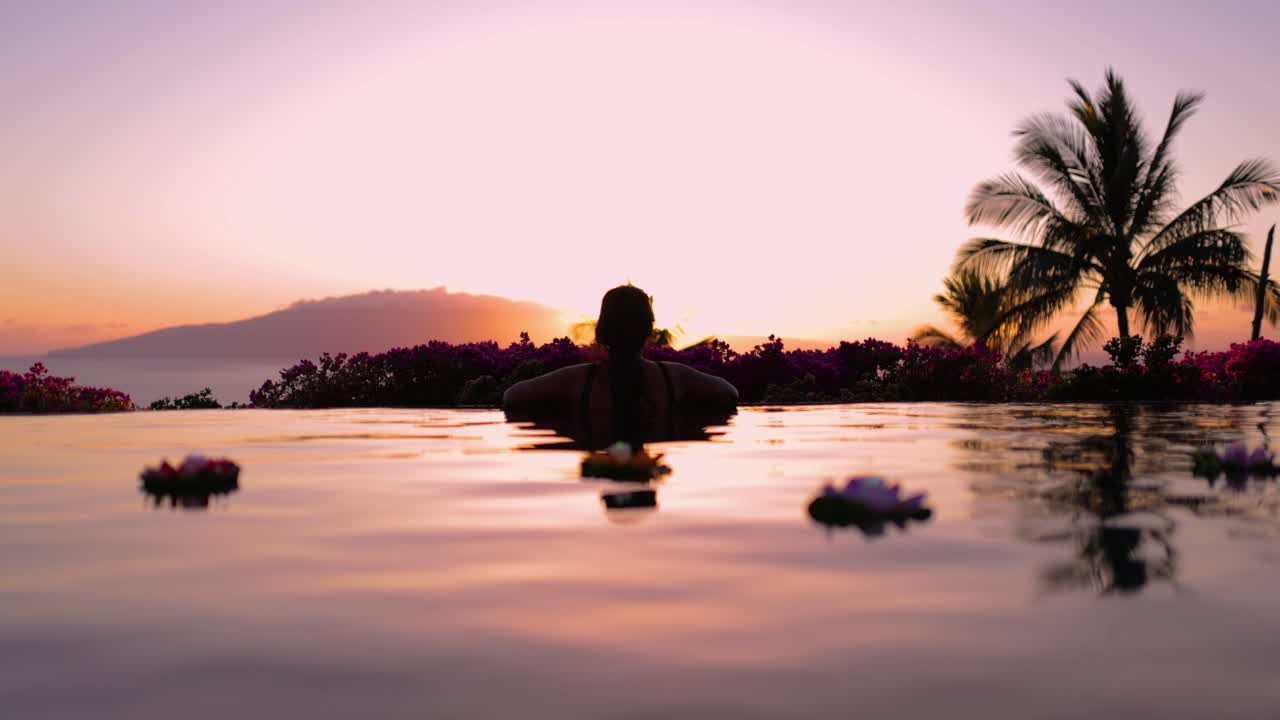 une jeune femme hawaïenne tatouée regarde le coucher du soleil derrière une île depuis une piscine à débordement