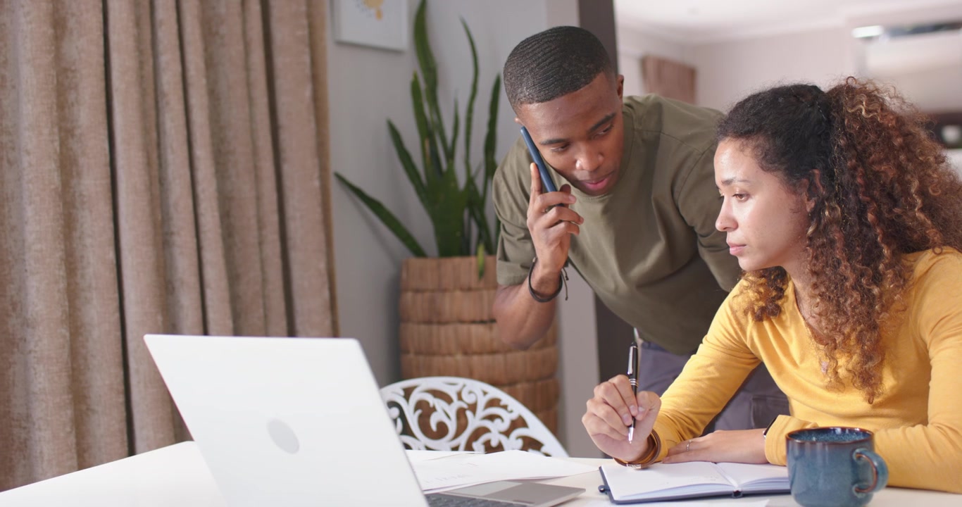 Couple coordinating after call home table, man holding phone pointing at laptop while woman noting