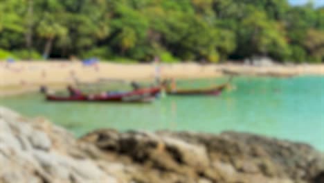 blurred view of a tranquil beach in phuket with turquoise waters, lush greenery, and traditional boats under bright daylight