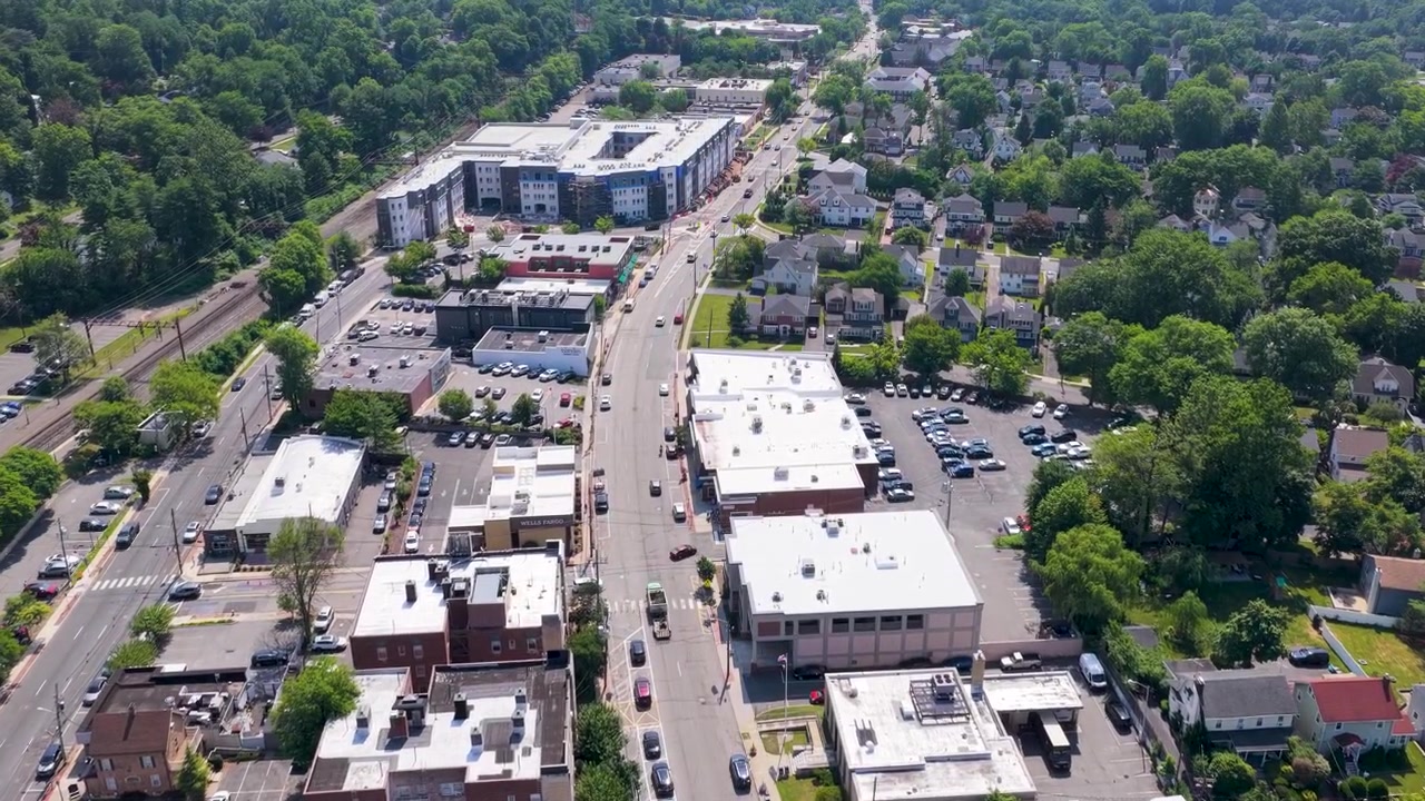 High angle drone footage capturing the bustling downtown district of millburn, new jersey, showing millburn avenue with its shops, apartment buildings, and steady traffic flow on a sunny day