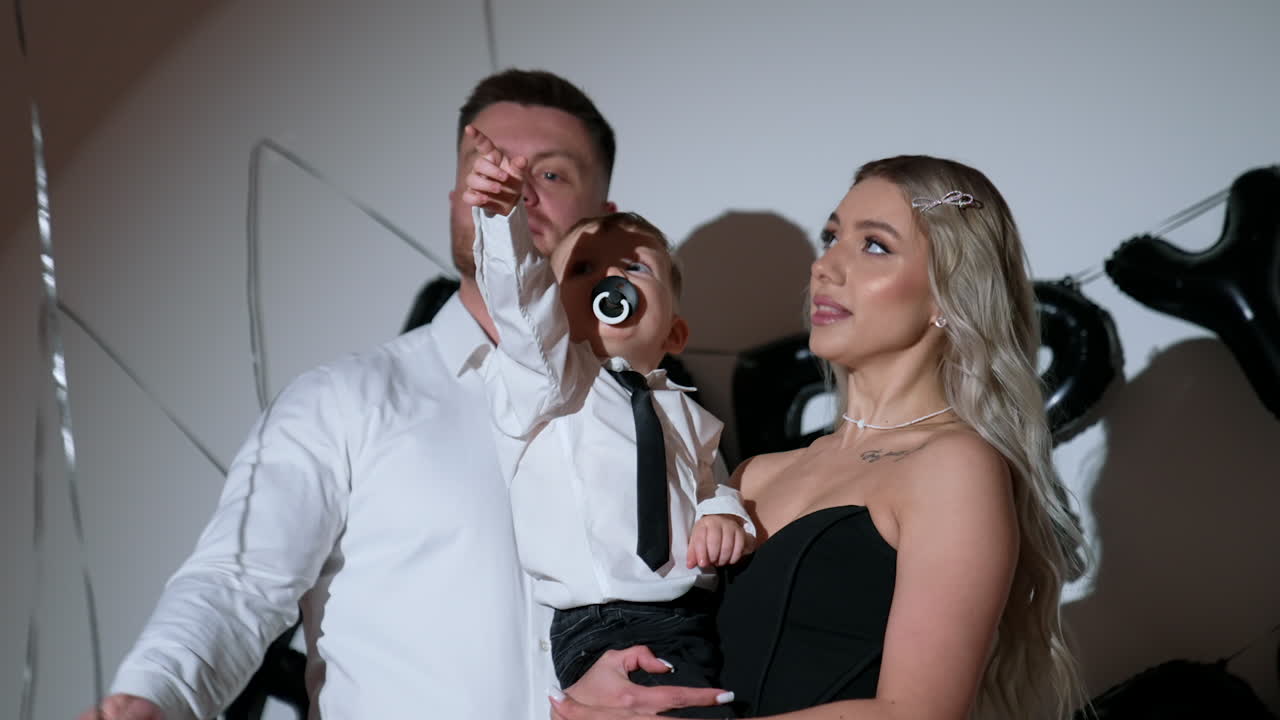 Loving Caucasian parents with their beloved baby in studio. Little kid sucking a pacifier pulls the balloon string.