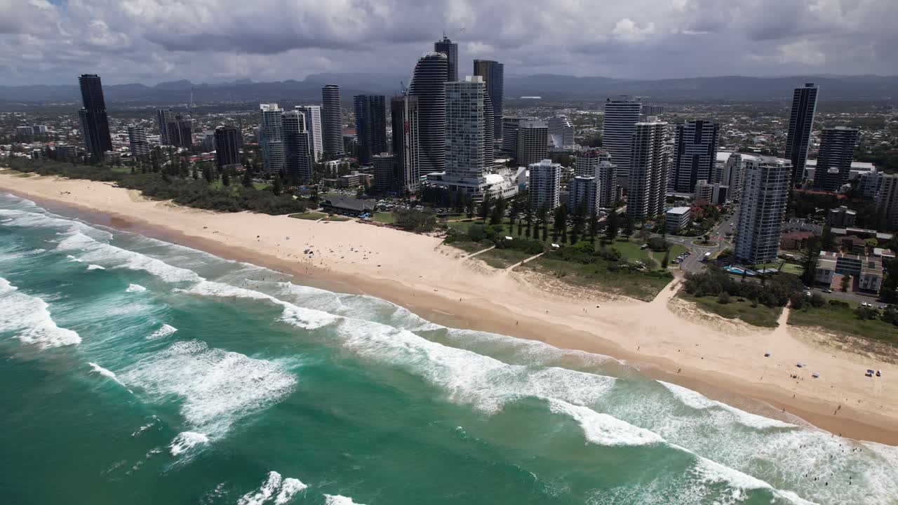 Waves Roll Onto the Shore Beside the Striking Jewel Residences and The Langham, Set Against a Backdrop of Modern High-rises in Gold Coast, Queensland, Australia - Orbit Drone Shot