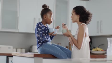 happy african american mother and daughter eating breakfast in kitchen, slow motion