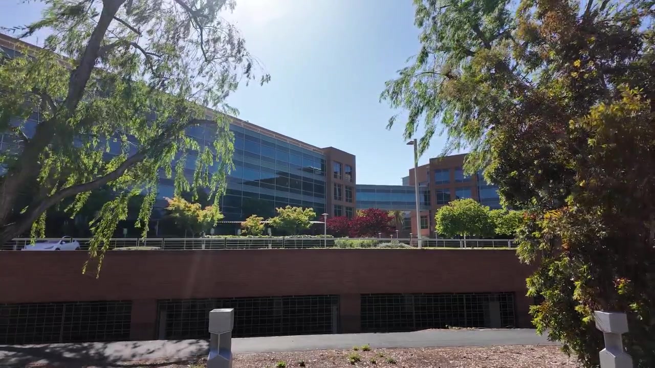 Moderno edificio de oficinas Googleplex en el campus de la empresa tecnológica en Palo Alto, California, con árboles verdes y un cielo azul brillante y soleado, mostrando innovación y éxito empresarial.