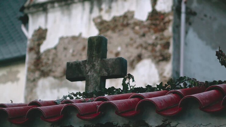 Una croce di pietra logorata dalle intemperie dietro un muro coperto di tegole rosse.