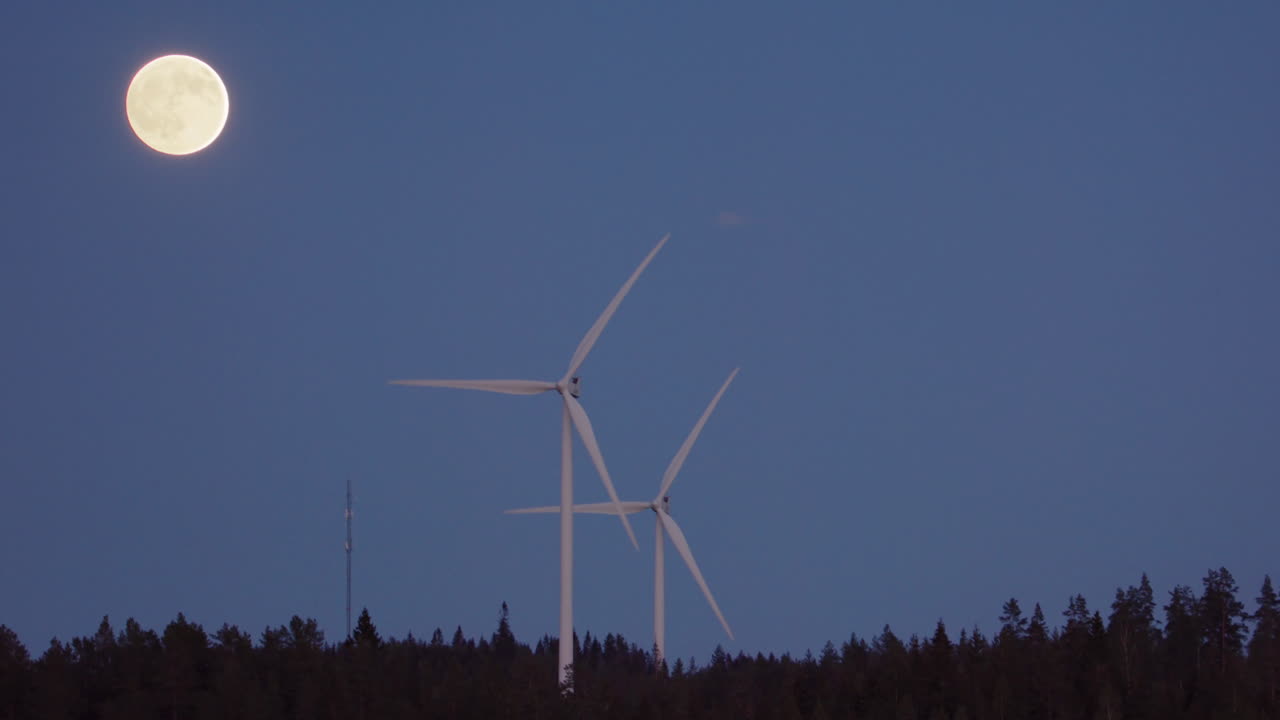 WIDE, SLOW MOTION, ZOOM IN - Turbines spin next to a rising full moon