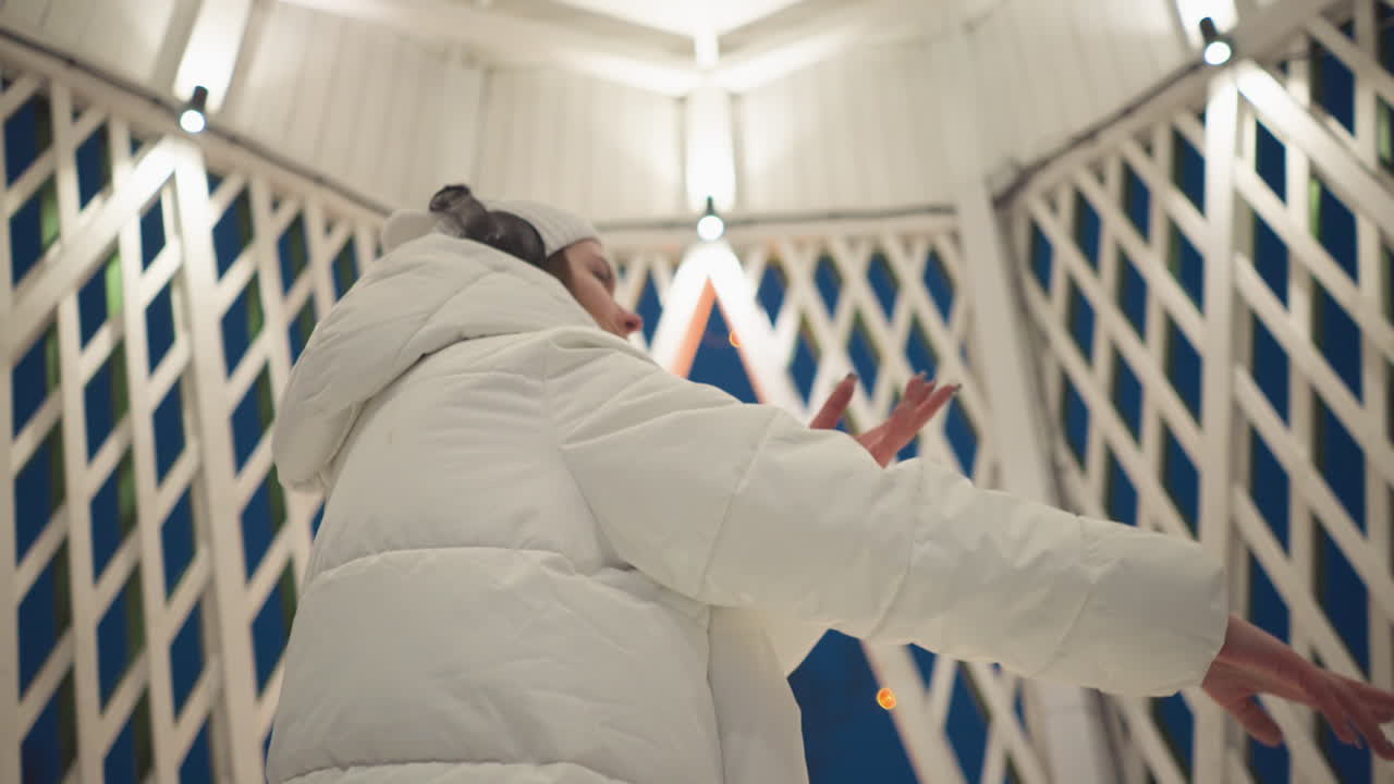 Student wearing white puffer coat and headphones dances inside latticed summer house adorned with glowing bulbs under warm overhead lights with graceful hand movements against deep blue sky backdrop