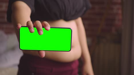 fat woman stands on huge stomach and shows phone with green screen. closeup of phone with chromakey. concept of excess weight. mockup