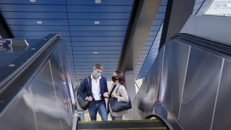 hombre de negocios y mujer de negocios montando escaleras mecánicas en la estación de tren usando máscaras faciales de ppe en la pandemia