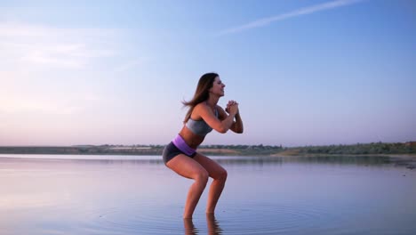 Side-view-of-young,-long-haired-woman-in-sportswear-doing-sit-ups-standing-in-the-water.-Happy,-healthy-lifestyle.-Beautiful-clear-blue-sky