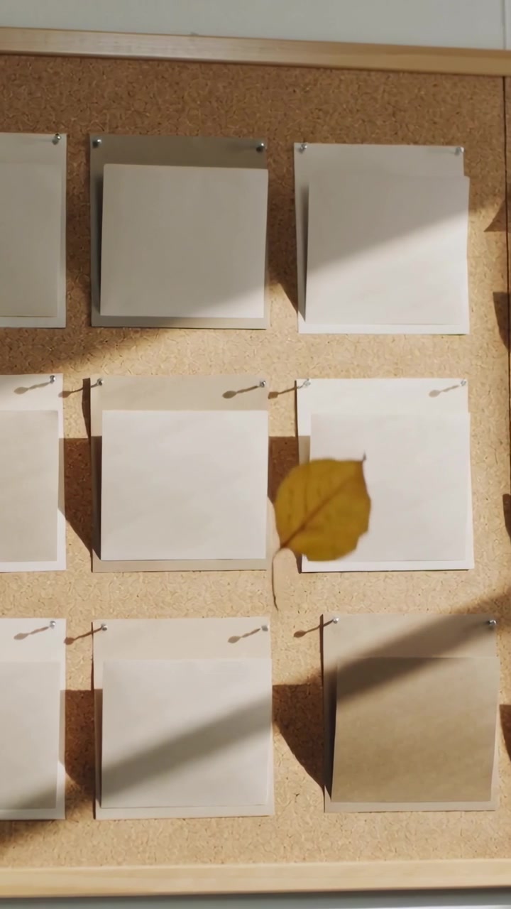 Vídeo vertical: Folha amarela se soltando, flutuando por blocos de notas, alfinetes em quadro de cortiça, sombras cruzando.