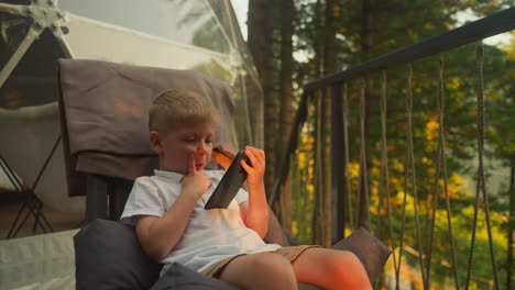 concentrated boy watches video on mobile phone enjoying vacation of glamping program. calm child relaxes receiving positive energy from nature slow motion