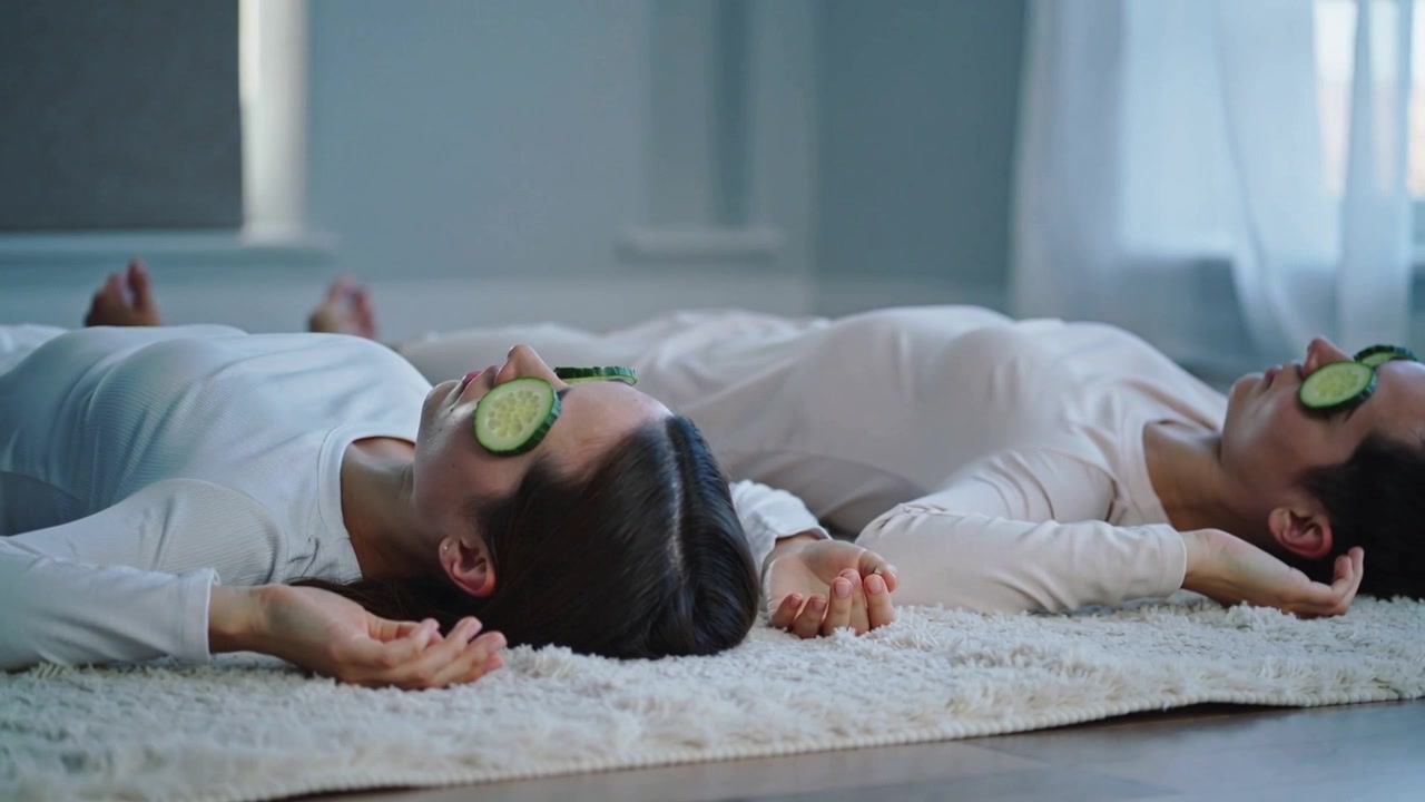 Placing cucumber slices, lying women in light loungewear adjusting on rug by window for skincare