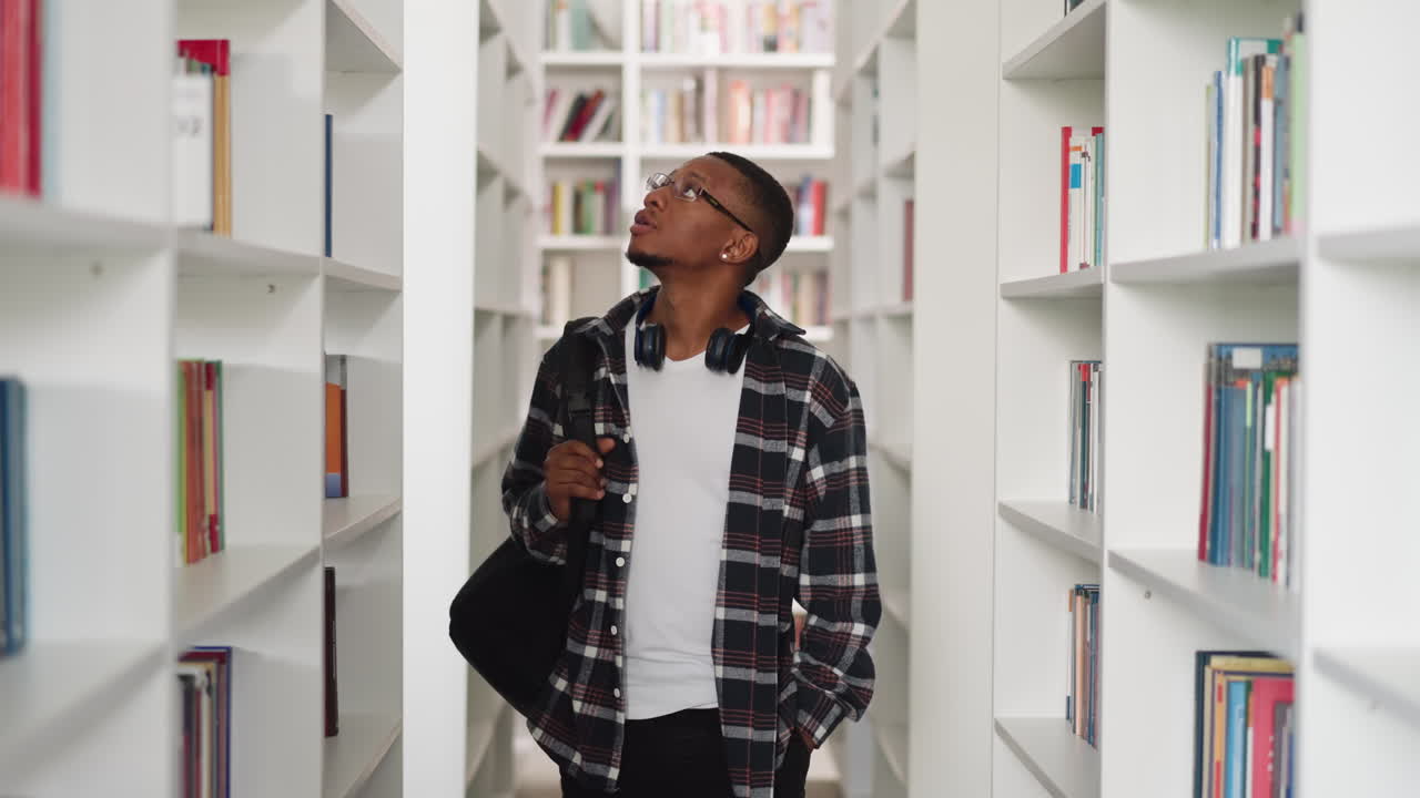 Young student looks around in college library. African American man in checkered shirt seeks textbooks in store. Informational sources storage
