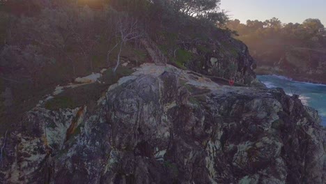 Rising-aerial-shot-of-a-cliff-with-a-man-sitting-at-sunset