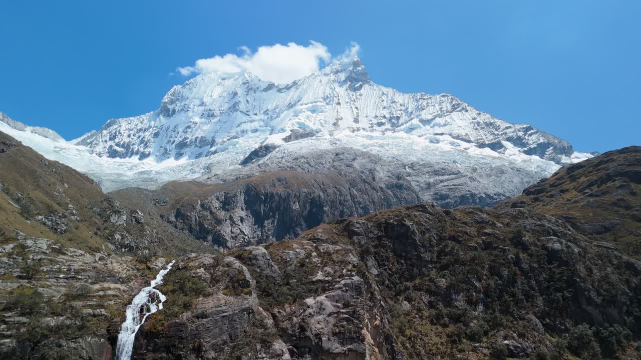 Stunning aerial shot of a glacier-fed waterfall plunging from Chacraraju mountain, feeding Laguna 69 and surrounded by the dramatic peaks of the Cordillera Blanca in Peru