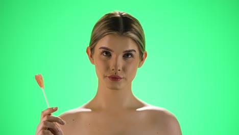 Static-slot-motion-shot-of-a-young-pretty-model-holding-a-heart-shaped-lollipop-in-her-hand-and-turning-it-back-and-forth-in-her-hand-while-looking-neutrally-at-the-camera-in-front-of-green-background