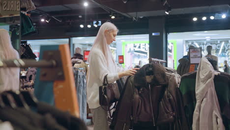 fashion lover with long silky hair and black handbag browses winter jacket rack inside transparent boutique window with shoppers walking through brightly lit modern mall environment