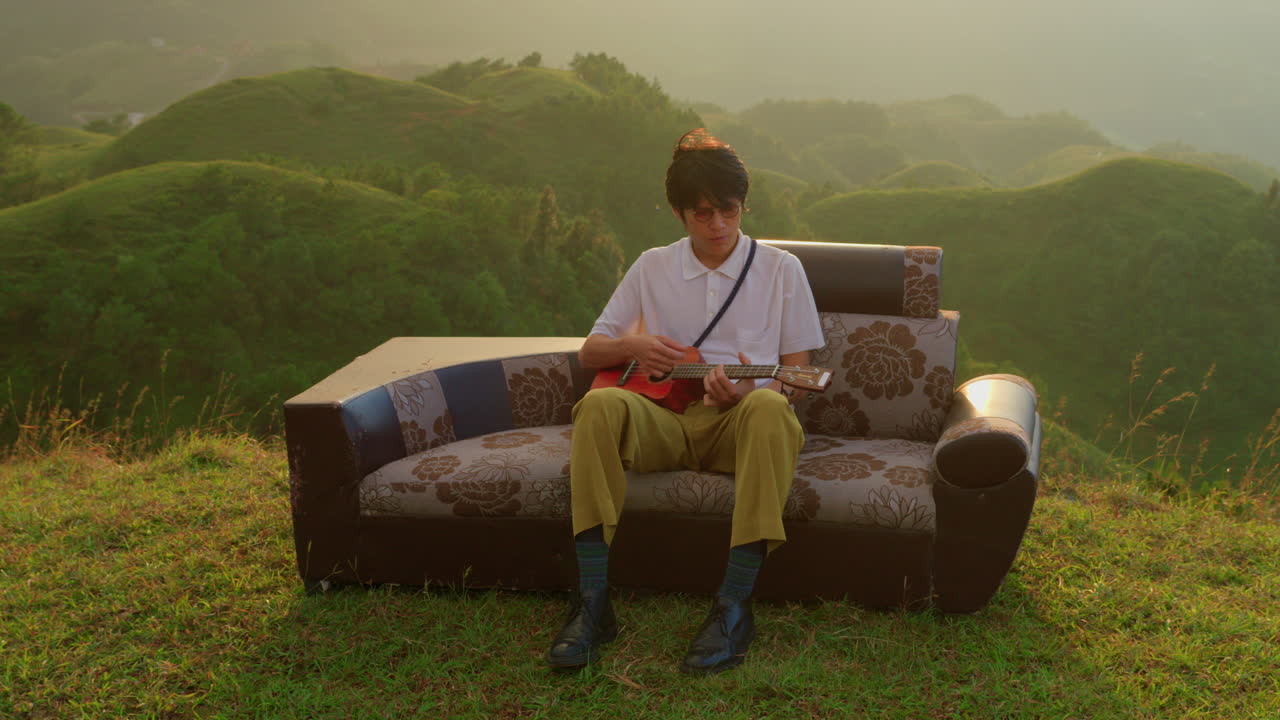 Man Plays Ukulele on Sofa Overlooking Mountains at Sunset