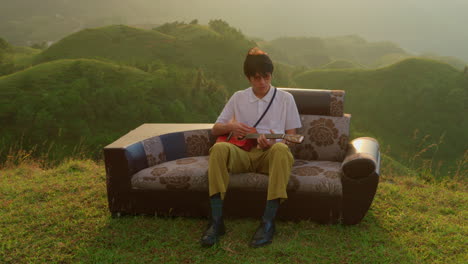 man plays ukulele on sofa overlooking mountains at sunset