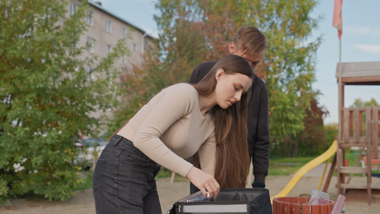 Młoda biała kobieta rozpakowuje torbę z dostawą, mężczyzna obserwuje w pobliżu placu zabaw, w tle budynek mieszkalny, rozpakowywanie zapakowanej żywności i artykułów spożywczych z izolowanej torby, sprawdzanie poprawności zamówienia