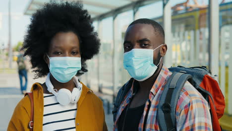 retrato de una pareja de jóvenes viajeros afroamericanos con mochila y máscara médica mirando la cámara al aire libre