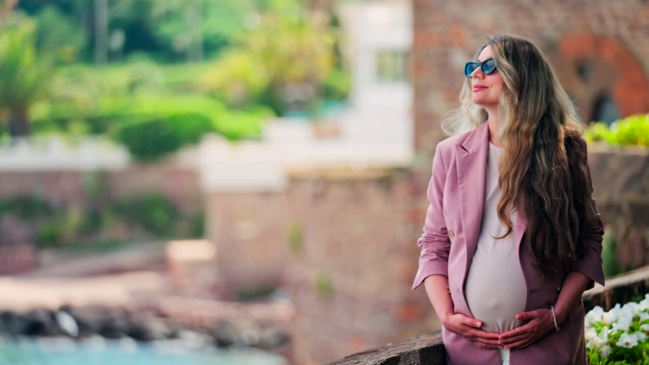 Pregnant woman with long hair, wearing sunglasses standing with the Chateau de la Napoule Castle in Mandelieu-La Napoule, France in the background