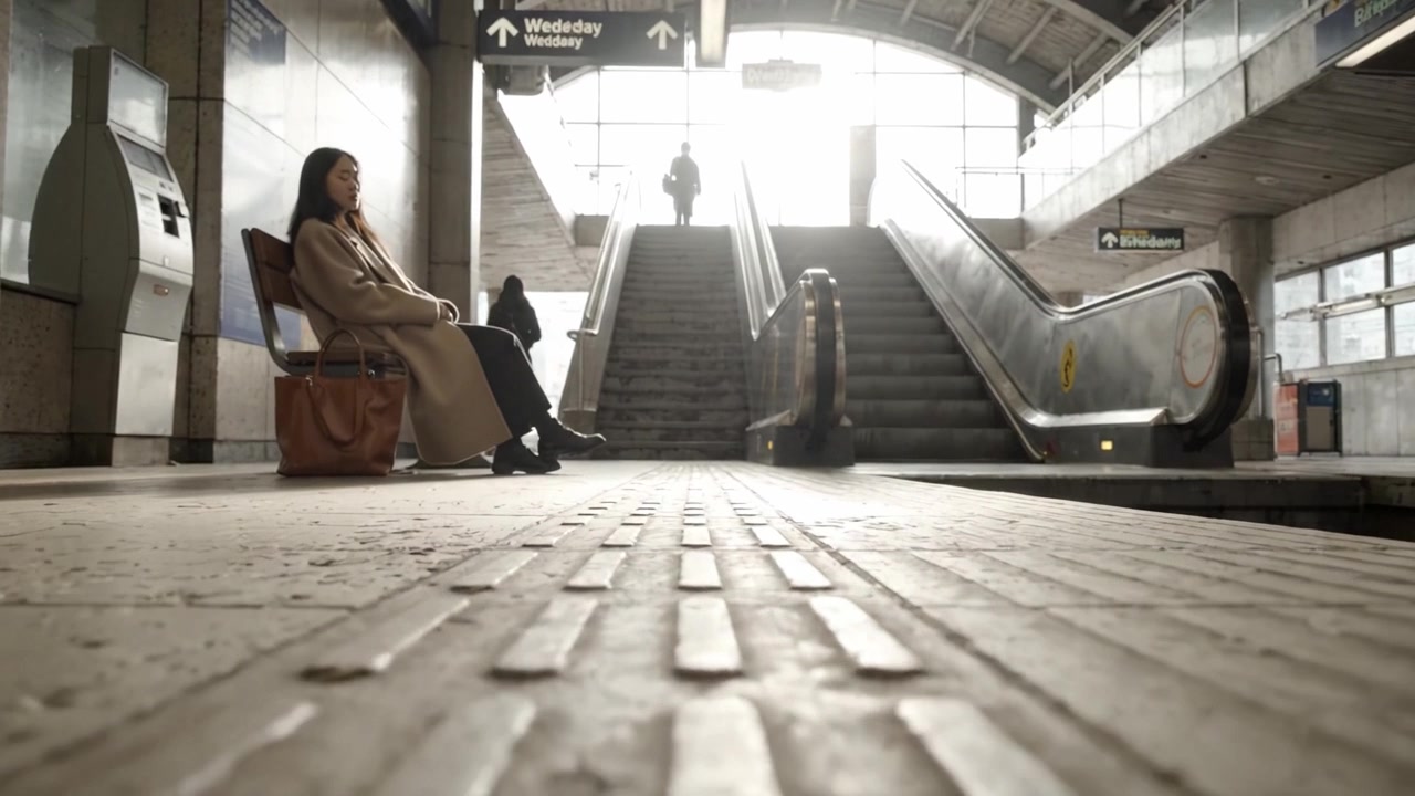Un momento di quiete alla stazione: una persona aspetta mentre un'altra si avvicina dalla scalinata, evidenziando la tranquillità quotidiana della vita urbana e del pendolarismo.
