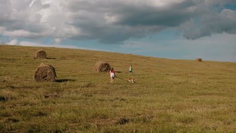 little boy in green top runs ahead through open countryside while his sister in pink follows, holding a dog on a leash, rolling farmland, scattered hay bales, and a blue sky with clouds