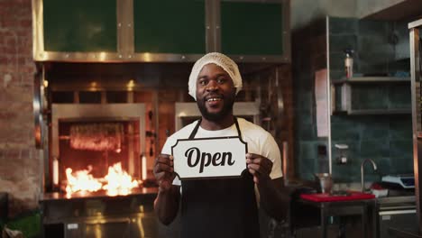 A-Black-person-in-a-special-headdress-poses-with-an-Open-sign-against-the-background-of-a-barbecue-in-a-doner-market