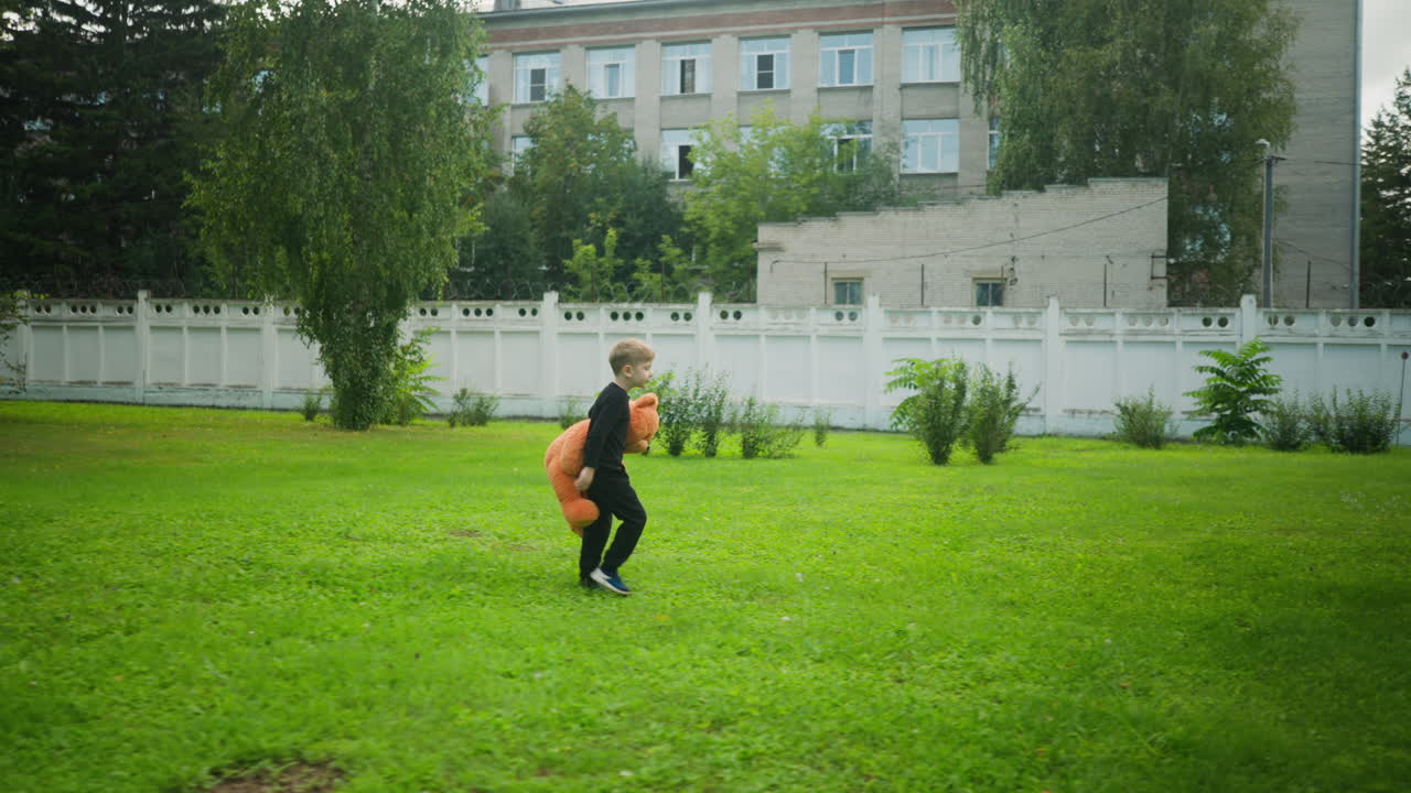 Little boy dressed in black outfit walking alone on green grassy field holding large teddy bear, approaching and jumping over small ditch near white fence with trees and building in background
