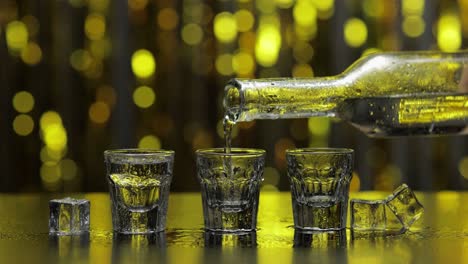 barman pour frozen vodka from bottle into shot glass. ice cubes against shiny gold party background