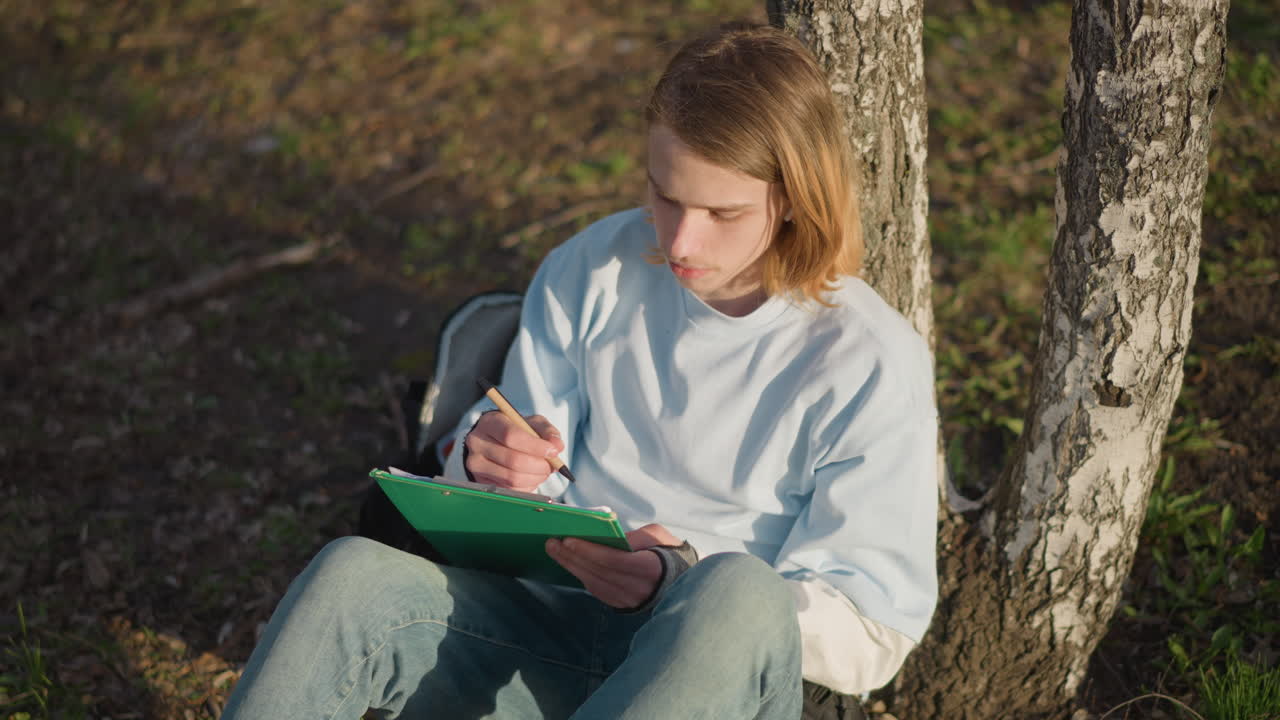 Молодой человек изучает папку на улице, подросток разбирает документы рядом с освещенным деревом, юный студент внимательно просматривает и сортирует файлы на открытом воздухе возле березы, сосредоточенно глядя на них.