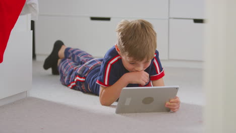 boy at home in bedroom wearing pyjamas watching movie or show on digital tablet lying on floor