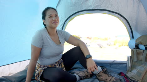 Smiling woman sitting in tent, wearing hiking boots and smartwatch, enjoying nature
