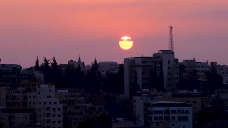 Lapso-De-Tiempo-De-La-Ciudad-Capital-De-Amman-En-Jordania,-El-Sol-Se-Sumerge-Detrás-De-La-Vista-Del-Paisaje-Urbano-Urbano-Con-Un-Hermoso-Cielo-Rosa-Al-Atardecer