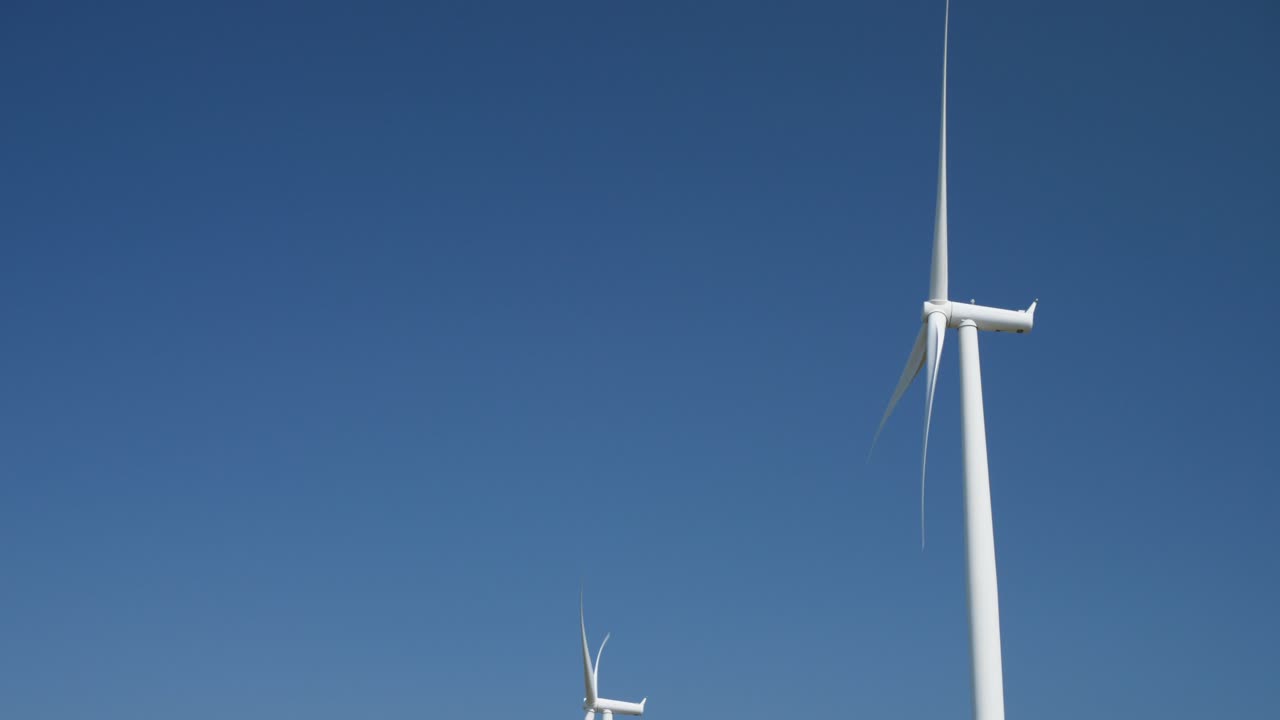 Wind turbines spinning under clear blue sky with minimal cloud coverage. Renewable energy sources generate clean electricity in vast open space. Sustainable technology in action