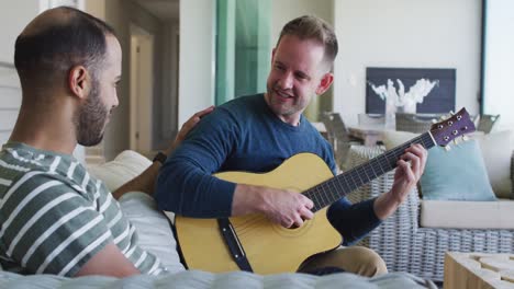 Multi-ethnic-gay-male-couple-sitting-on-couch-one-playing-guitar