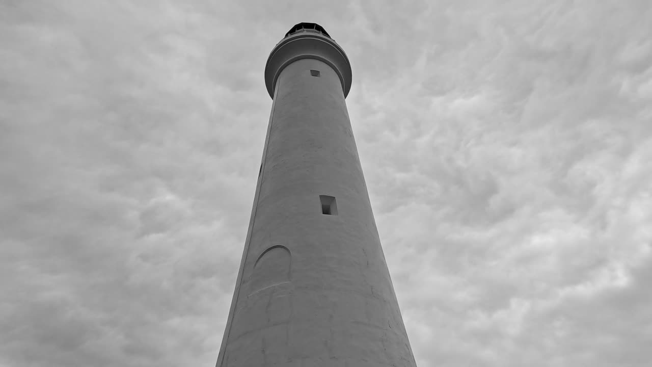 Low-angle camera pans upward along a tall white lighthouse against dramatic, cloudy skies. Overcast natural lighting, smooth vertical movement, moody coastal atmosphere