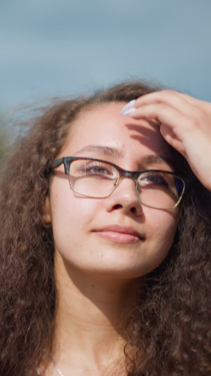 mujer con cabello rizado y gafas caminando al aire libre con una cálida sonrisa, ajustando el cabello hacia atrás bajo la luz del sol, con edificios borrosos y vegetación en el fondo