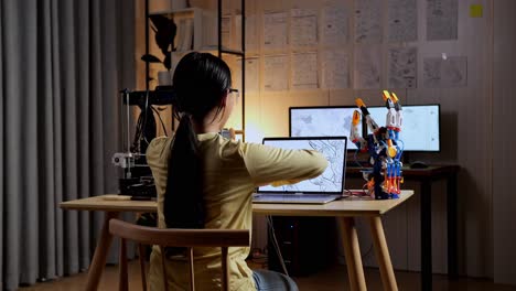 back view of teen asian girl with 3d printing stretching while designing a cyborg hand on a laptop at home