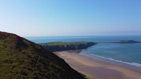 descending aerial view of rhossili bay in gower peninsula with blue sea waves and cliffs background and moorland in foreground 4k