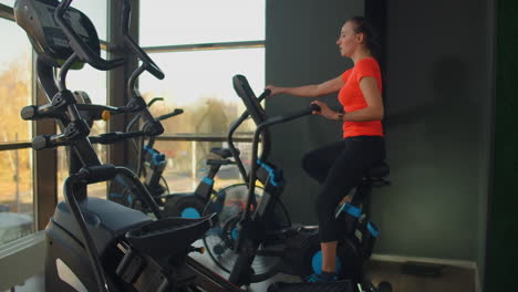 Young-active-woman-spinning-a-air-bike-in-gym-with-trainers.-Female-training-on-air-bike